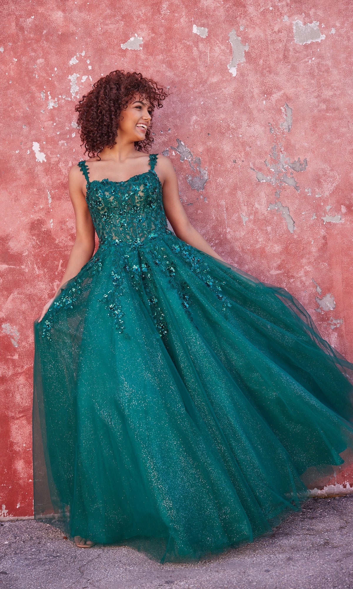 A woman in the Ellie Wilde A-Line Long Designer Ball Gown EW35123 poses against a weathered pink wall, holding the sides of her sparkly green formal dress and smiling.